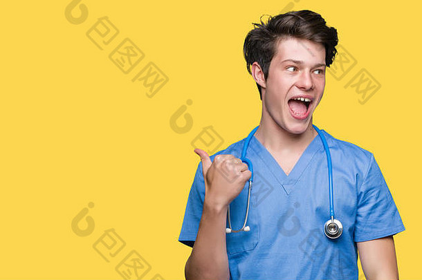 年轻的医生穿着隔离背景的医疗制服，面带笑容，大拇指朝上指向侧面。