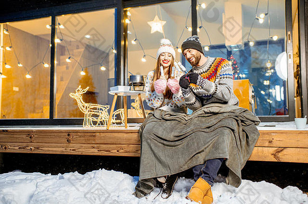 寒假期间，一对年轻夫妇坐在山上现代住宅的露台上，穿着格子布和棉花糖热身