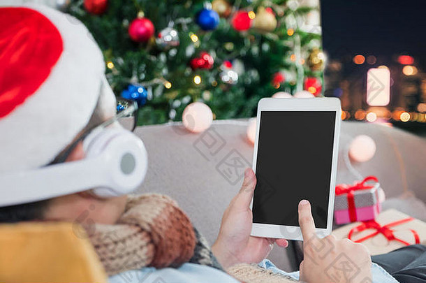 男人在平板电脑上看新年视频，快乐的人们躺在沙发上，晚上从家里抱着圣诞树倒计时，庆祝节日