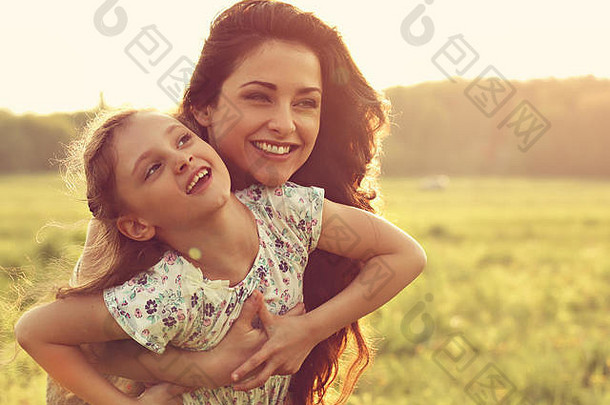 在夕阳明媚的夏日背景下，快乐的母亲与她轻松快乐的小女孩拥抱在一起。爱情的特写彩色肖像。
