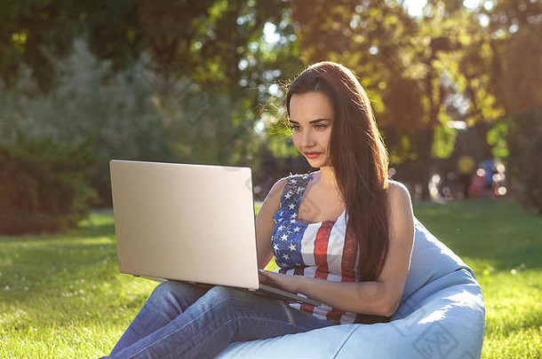 年轻可爱的女孩拿着笔记本电脑，坐在花<strong>园</strong>或公<strong>园</strong>的豆包上，绿草丛中。网上购物概念