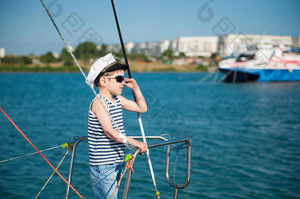 夏日旅行期间，一个戴着白色船长帽和条纹背心的快乐小男孩站在游艇甲板上调整着海港的太阳镜