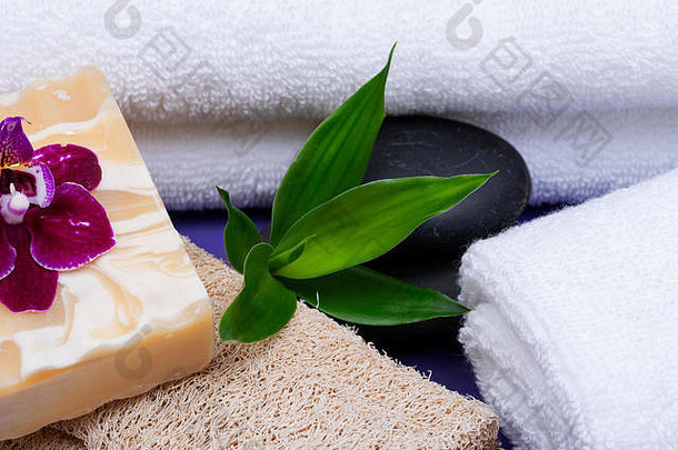 Spa健康概念。天然丝瓜海绵、杏仁山羊奶皂、白色毛巾、玄武岩石头、紫色背景上的竹子和兰花。