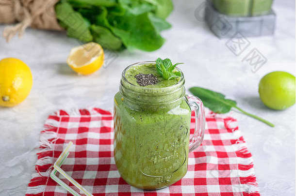 健康的绿色菠菜冰沙放在一个罐子里，杯子上装饰着薄荷和芝麻籽，配料放在白色大理石桌上的方格餐巾上。选择