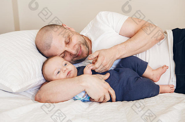 中年白人父亲穿着白色t恤躺在床上，新生的儿子握着他的手和手指，养育孩子