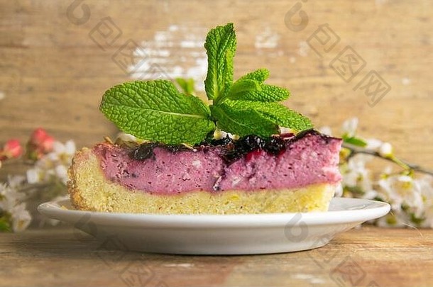 木桌上放着几片蓝莓<strong>慕斯</strong>蛋糕，上面有镜面釉装饰的浆果和薄荷叶，白色的盘子上有白色的花朵