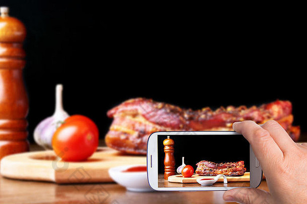 手持智能手机的男子在黑暗中的砧板上拍照烤排骨烧烤条纹里脊牛排加芝麻酱和西红柿