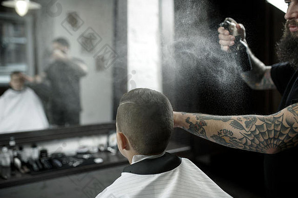 <strong>儿童</strong>理发师在黑暗的背景下给小男孩理发。心满意足的可爱学龄前男孩正在理发。大师的手上有“剃须”这个字的纹身