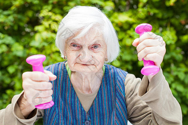 照片中，一位精力充沛的老妇人手持粉红色的哑铃，在花园里做户外运动