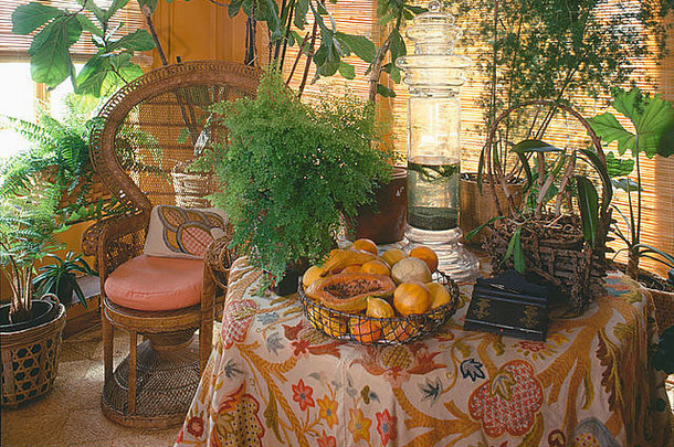 在七十年代的餐厅里，一碗水果和郁郁葱葱的绿色室内植物摆在藤制孔雀椅旁的带图案的桌布上