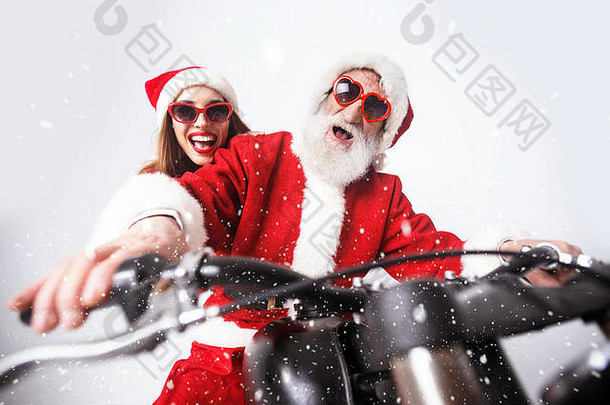 戴着白胡子的圣诞老人和戴着圣诞帽、红色毛衣和太阳镜的年轻老太太骑着摩托车高速行驶，新年、圣诞节、节假日、纪念品、礼品、<strong>购</strong>物、折扣、商店、雪人圣诞老人、化妆、发型、<strong>狂欢</strong>节。