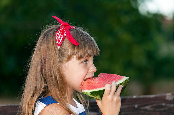 一个头发上系着红丝带的美丽小女孩的肖像。她吃着西瓜，享受着美丽的夏日。玩耍、成长、快乐