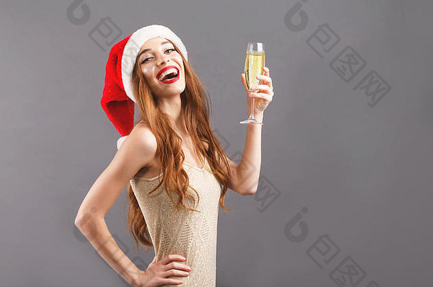戴着红色圣诞老人帽、红唇站在灰色背景上的可爱的长发女人，她说祝酒词，喝香槟，新年，圣诞节，假期，纪念品，礼物，<strong>购</strong>物，折扣，商店，雪人圣诞老人，化妆，发型，<strong>狂欢</strong>节。