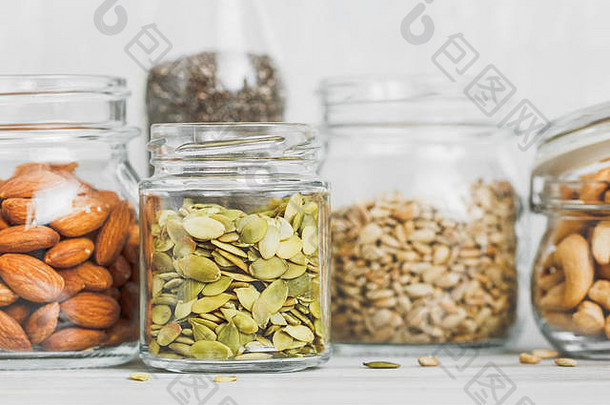 各种各样的坚果和种子放在玻璃罐里，放在白色背景下的白色木桌上。素食和有机食品的概念。一套照片。