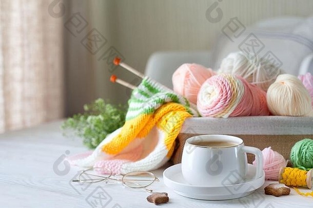 针织亮条纹织物。毛线和棉线放在篮子里。卡布奇诺咖啡、饼干和玻璃杯放在白色木质背景上。