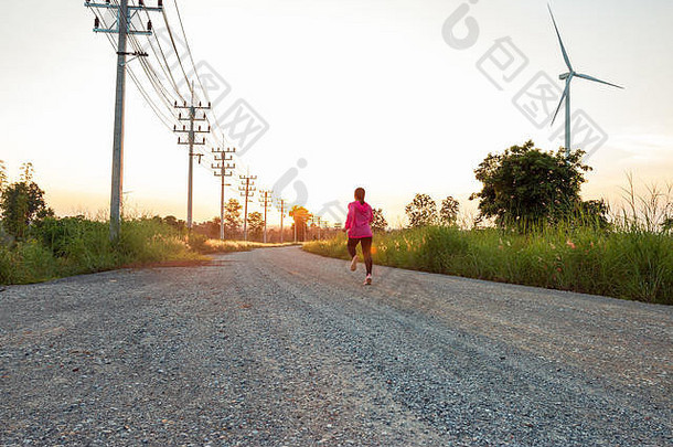傍晚日落时，该地区的风力涡轮机产生清洁能源电力，一名妇女正在路上慢跑。