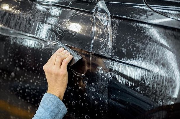 在详细车辆车间，工人用刮刀在车身上粘贴防碎石膜，特写。用特殊薄膜保护车身的概念