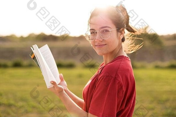 一位年轻女子一边在公园里微笑着欣赏夕阳的余晖，一边读着好书。
