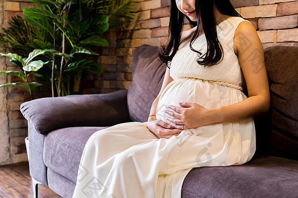 一位年轻的亚洲孕妇坐在家里的沙发上，抚摸并抱着一个肚子里的未出生的孩子