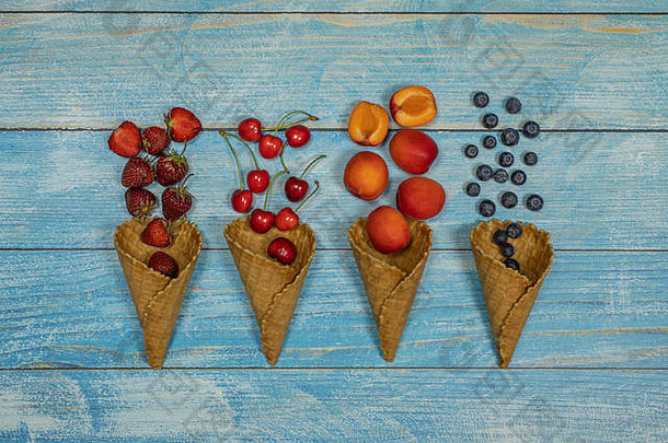 浆果和水果冰淇淋。在蓝色木质背景上，将各种新鲜水果、蓝莓、草莓、樱桃、杏平放在华夫格蛋卷中。夏季甜点菜单概念。冰淇淋制作