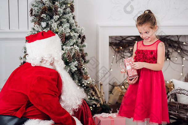 圣诞老人和孩子们在壁炉前打开礼物。孩子们的父亲穿着化装，留着胡子，打开圣诞礼物。小女孩帮着拿礼物袋。圣诞树下的一家人在背景上的火上。