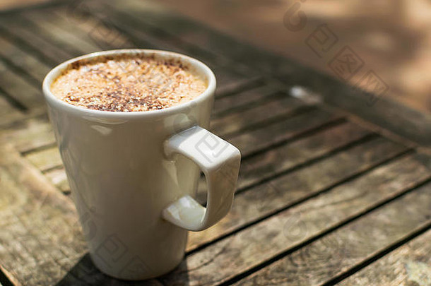 在英国的一家户外咖啡馆里，一杯卡布奇诺咖啡在阳光下放在一张木桌上，白色的杯子里。