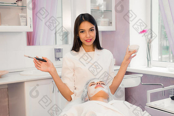 快乐的年轻的美容师应用面具女脸刷站微笑女人说谎