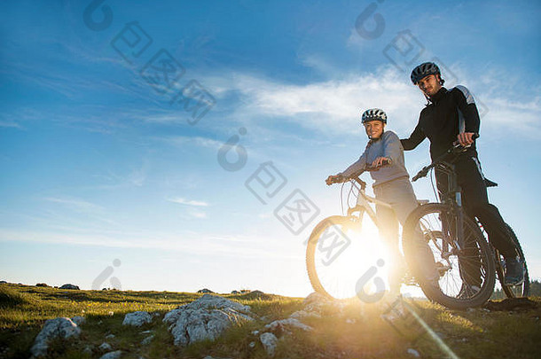 一对骑着山地自行车的夫妇站在夜空<strong>下</strong>的小山上，享受着日落时的灿烂阳光。