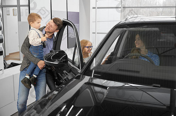 坐在新型昂贵黑色轿车驾驶座上的妇女。父亲牵着男孩的手，微笑着展示着汽车。快乐家庭在汽车中心展厅测试和观察汽车。
