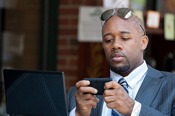 一位相貌英俊的非裔美国商人手持智能手机，在笔记本电脑或上网本上工作。