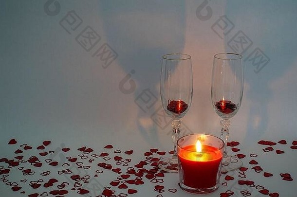 两个香槟酒杯，前面有一支轻蜡烛，周围是白色背景上的心形五彩纸屑，并留有复印空间。。