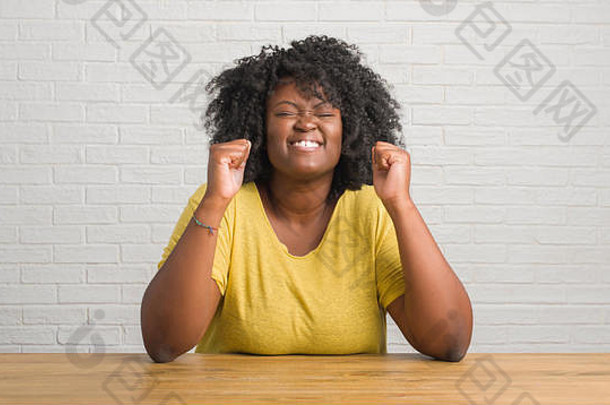 年轻的非洲裔美国妇女坐在家里的桌子上，举起双臂，闭上眼睛，兴奋地尖叫着，庆祝疯狂和疯狂的成功。Wi