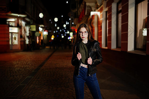 女模特戴眼镜、牛仔裤和皮夹克，背着背包，对着城市街道的夜景肖像。