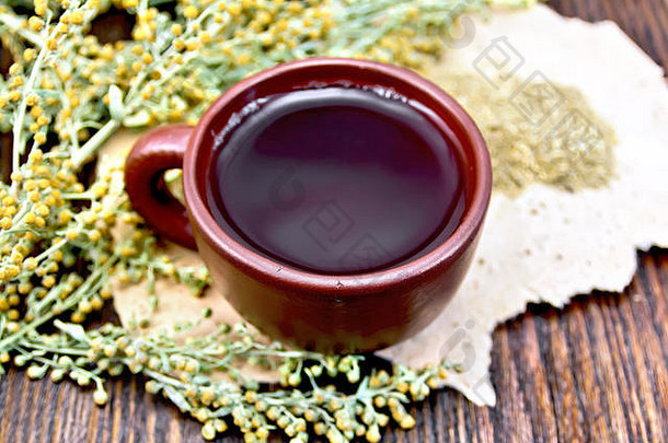 棕色粘土杯中的草药茶，灰色的山艾树枝，在深色木板上粗糙的纸上干燥的艾草