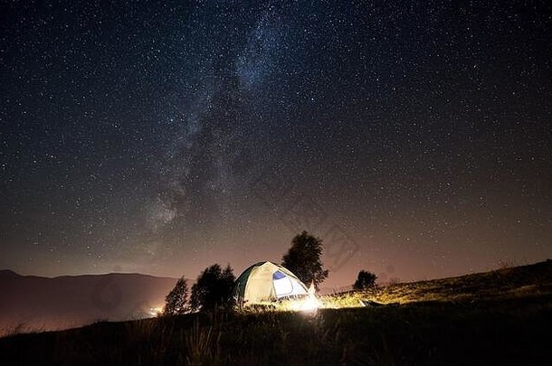 游客在夏夜露营。在充满星星和银河的神奇夜空<strong>下</strong>，照亮了帐篷和篝火。背景是美丽的<strong>星空</strong>、群山和发光的城镇