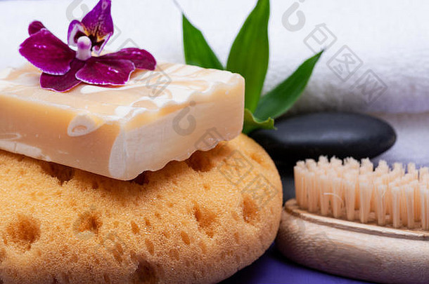 Spa健康概念。泡沫海绵、杏仁山羊奶皂、天然鬃毛木刷、玄武岩石头、紫色背景上的竹子和兰花。