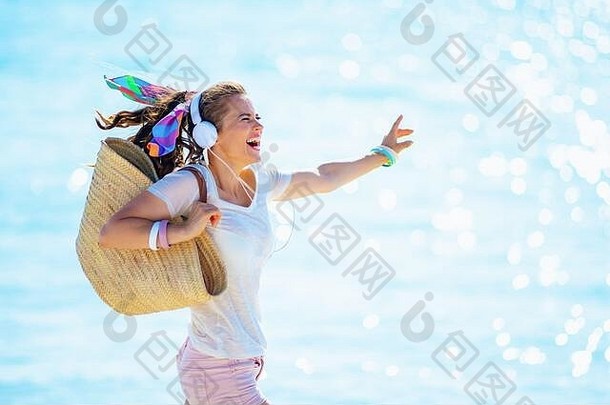 快乐的年轻的<strong>女人</strong>白色t恤粉红色的短裤海滩稻草袋听音乐耳机跳海洋海岸