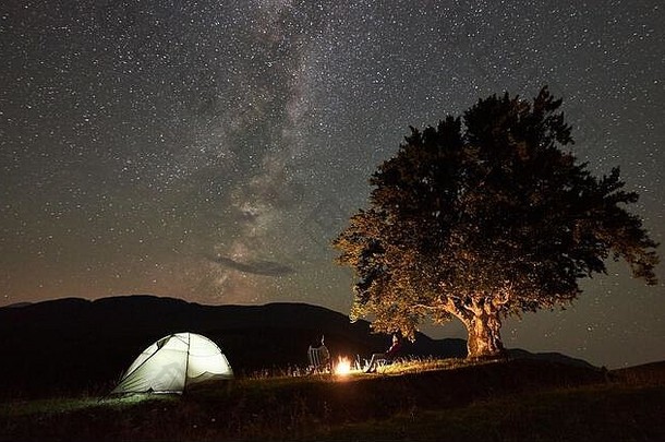 活跃的家庭游客在夏夜露营时休息。背景：男人和女人坐在篝火旁的椅子上，帐篷和大树，美丽的<strong>星空下</strong>布满了星星和银河。