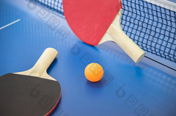 橙色的乒乓球和两个红黑相间的木制球拍，放在一张有格子的蓝色桌子上
