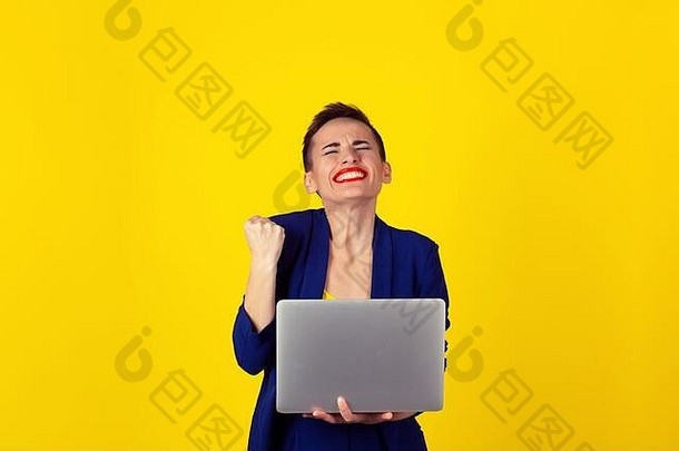网上彩票<strong>中奖</strong>者。拿着笔记本电脑挥舞拳头的女人开心地笑着。女孩短发红唇蓝色套装黄色衬衫隔离黄色背景墙