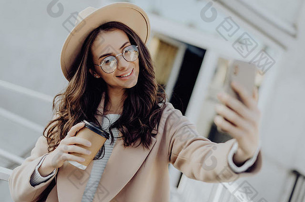 美丽的年轻的时尚的金发女郎女人穿外套走在户外采取自拍移动电话持有外卖咖啡杯