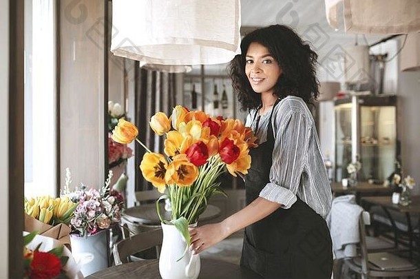 穿着围裙的美丽花商在花瓶里创造了一大束五颜六色的郁金香。年轻的非洲裔美国女孩，手持五颜六色的鲜花。肖像画