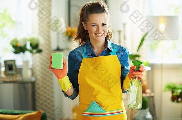 身着橙色围裙、粉色橡胶手套、带环保清洁用品喷雾瓶和绿色海绵h的微笑的中年家庭主妇肖像