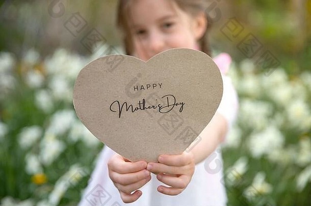 母亲节快乐写在一张心形卡片上，卡片的背景是花草，由一个女孩拿着