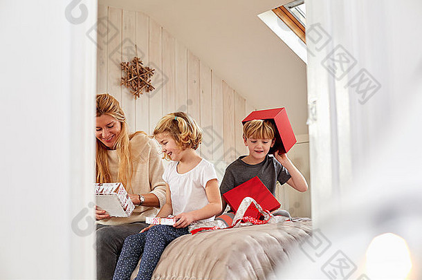 在一个家庭的圣诞节早晨。一位母亲和两个孩子坐在床上打开礼物。