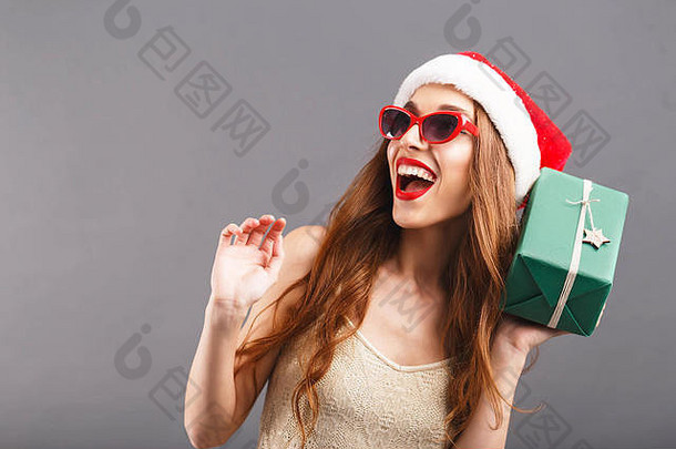 戴着红色圣诞老人帽子和红唇太阳镜的滑稽可爱的女人摇着绿色包装的礼物，她笑着站在灰色背景上，新年、圣诞节、假日、纪念品、礼物、<strong>购</strong>物、折扣、商店、雪人圣诞老人、化妆、发型、<strong>狂欢</strong>节。