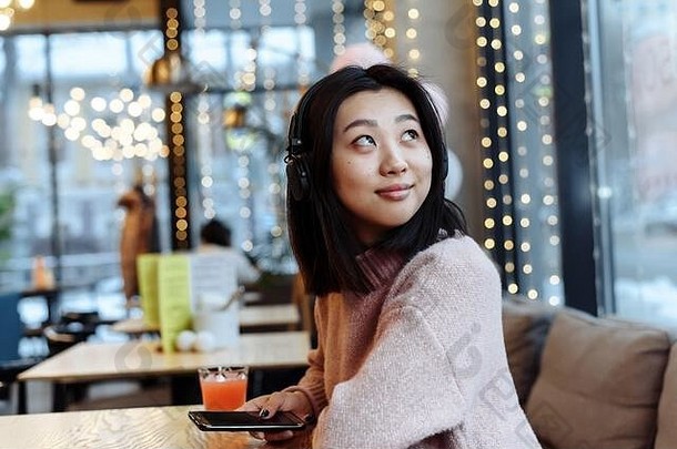 一个可爱的亚洲女孩坐在咖啡馆里，戴着大蓝牙耳机听音乐，喝着<strong>鲜榨果<strong>汁</strong></strong>。