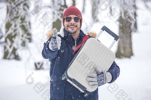 雪地上年轻时尚男士的肖像，手持手提箱，竖起大拇指。寒假旅游概念。