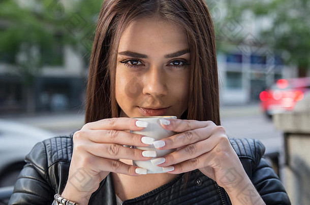 一个棕色头发的白人女孩在街上的露台上喝<strong>咖啡</strong>的照片。她用双手捧着<strong>咖啡</strong>杯。