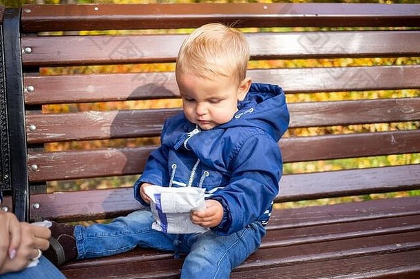 婴儿外部卫生和洗手理念。穿着蓝色衣服的可爱的小婴儿坐在公园的长椅上，手里拿着一包餐巾和一只手